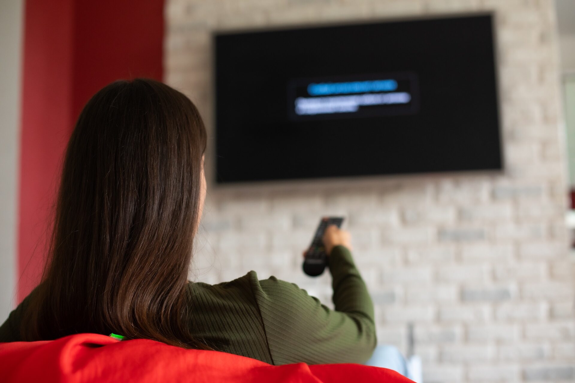 Back view photo of brunette young woman in casual clothes sitting on couch in living room, holding remote, switching channels on TV, weekend at home, rest and relax