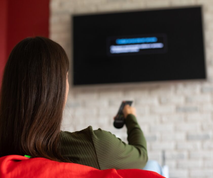 Back view photo of brunette young woman in casual clothes sitting on couch in living room, holding remote, switching channels on TV, weekend at home, rest and relax